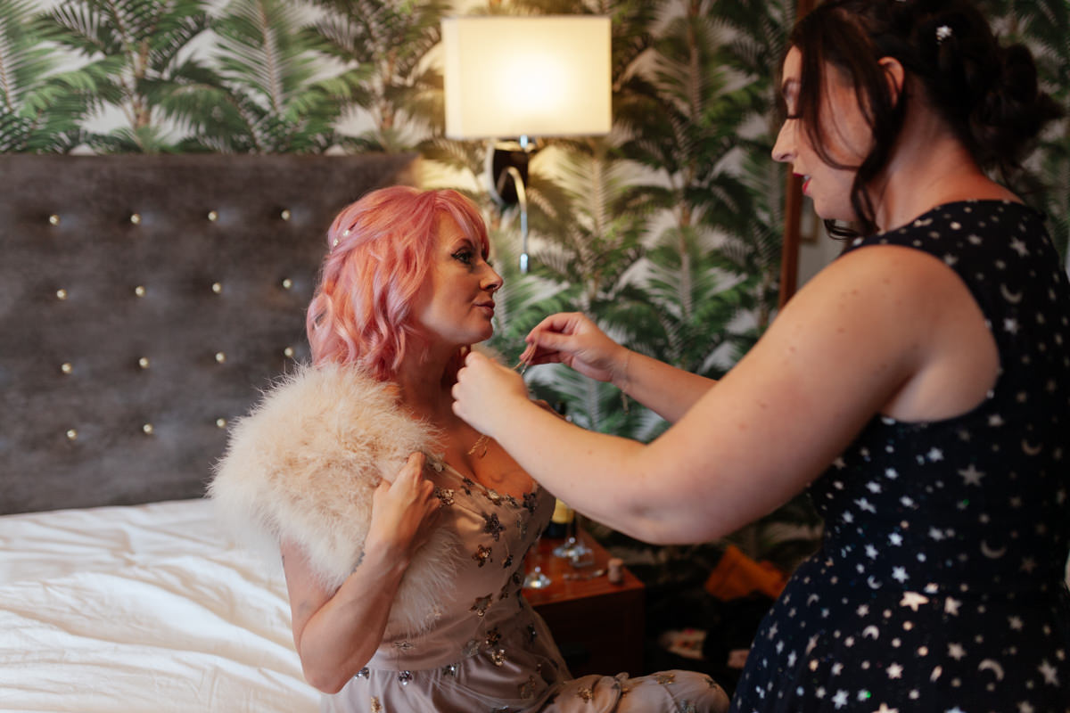 maid of honour helps bride put on her necklace whist she is sitting on a bed