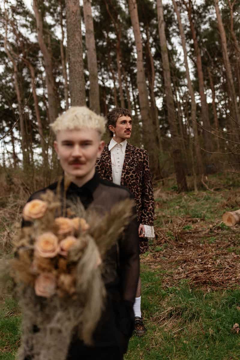 Person in a sheer black top holding dried flowers, with another person in a patterned coat standing among tall trees.