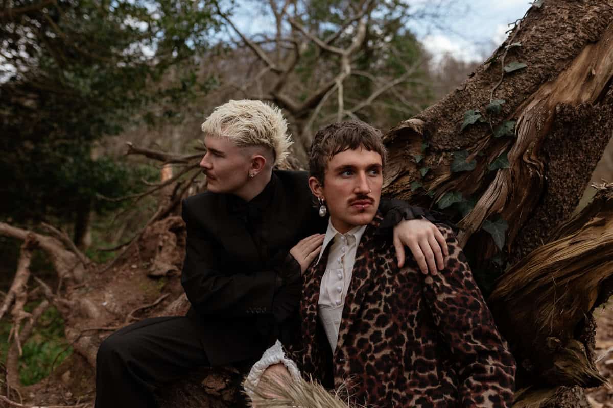 Two people sitting together on a fallen tree in a forest, one in a black suit and one in a leopard‑print coat.