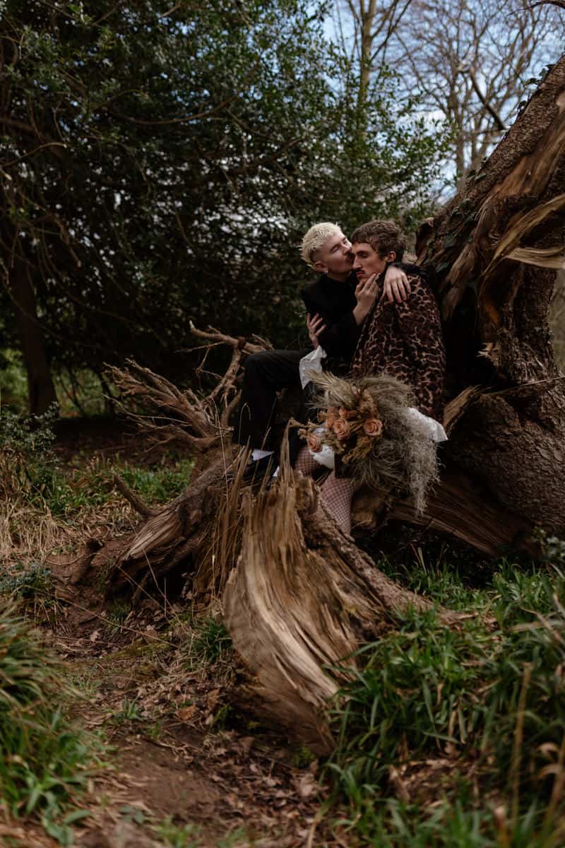 Two people sitting close together on a fallen tree, one holding dried flowers, sharing an intimate moment in the woods.