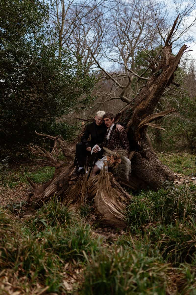 Two people sitting on the roots of a fallen tree, one holding a dried‑flower bouquet, surrounded by forest.