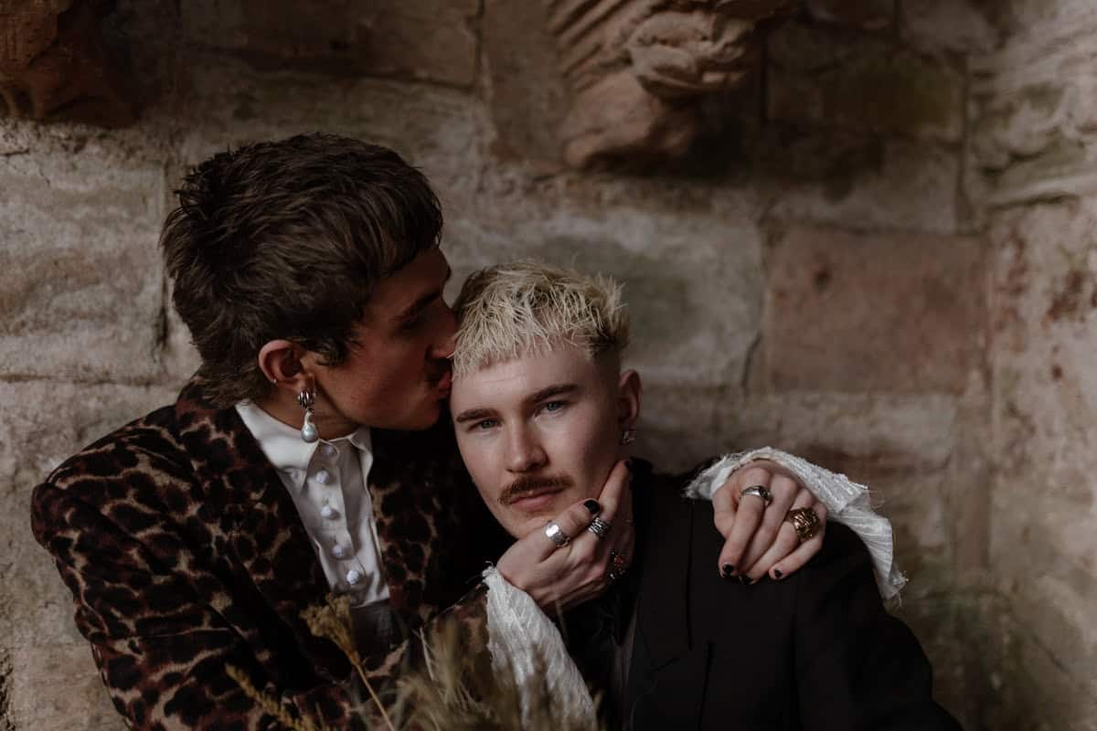 two grooms embrace holding each other, one stares into the camera while the other cups his face and softly kisses his forehead. Thrive conference shoot session with Esme whiteside at Dunglass estate