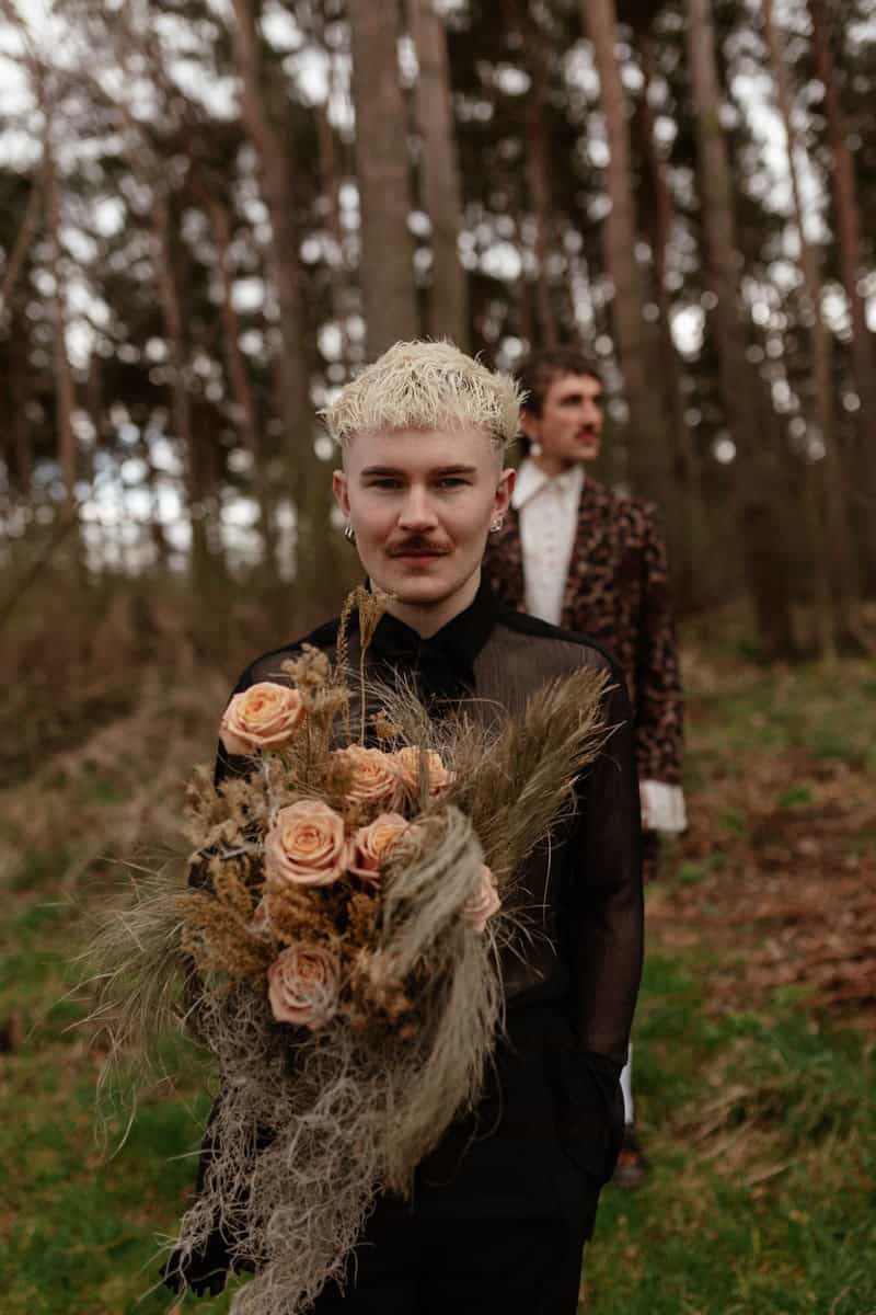 Person in a sheer black top holding a large dried‑flower bouquet, standing in a forest with another person behind.
