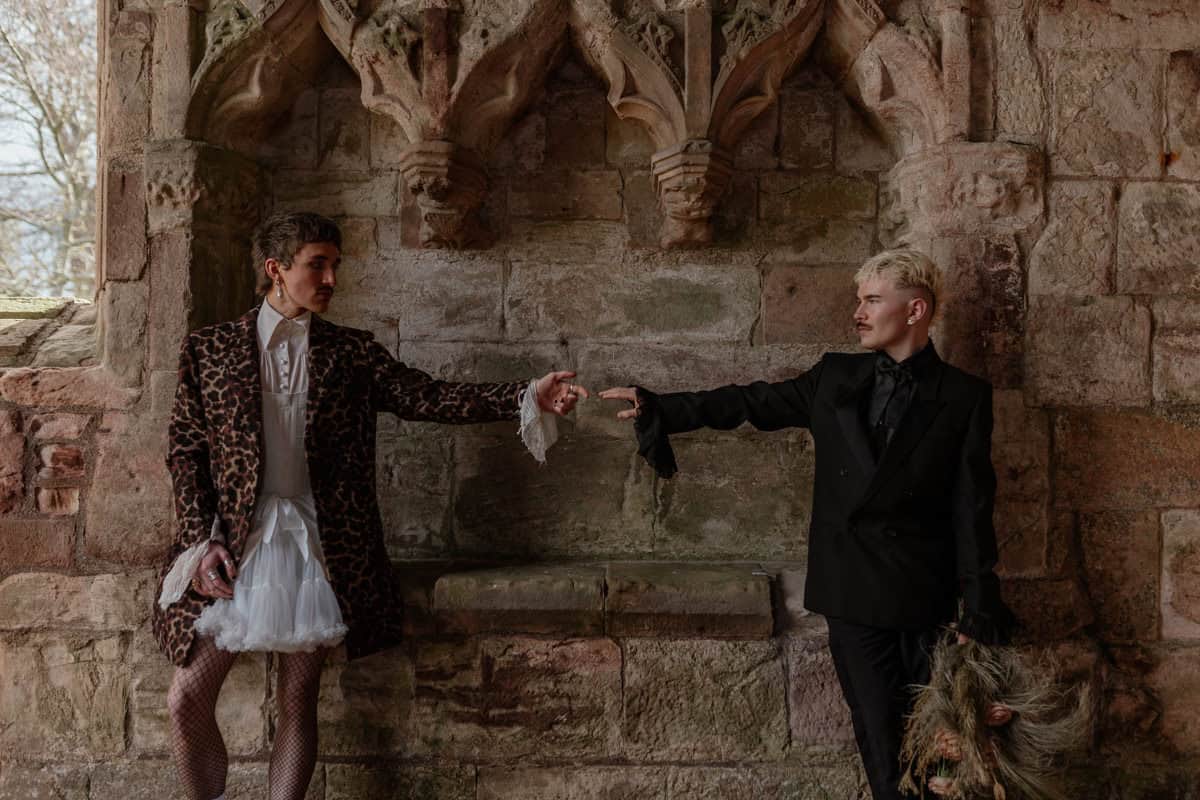 Two people reaching toward each other before carved stone arches, one holding dried plants, in a dramatic historic setting.