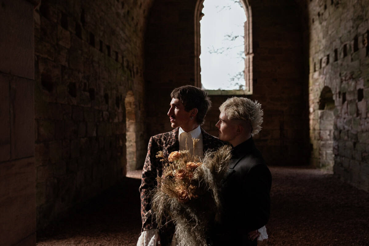 Two people standing in a dim stone interior, one holding a bouquet, light falling through an arched window behind them.
