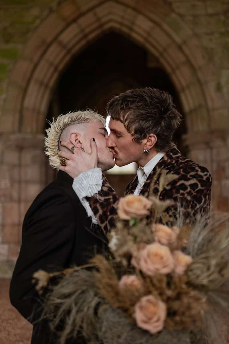 Two people kissing inside Dunglass Collegiate Church, framed by a stone arch and holding soft peach‑toned flowers.