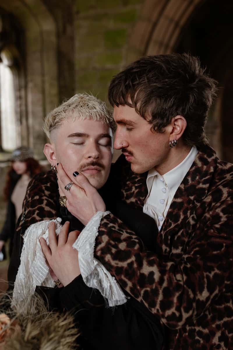 Two people in ornate outfits sharing an intimate moment against a stone wall in a historic setting.