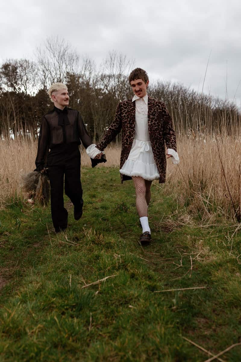 Two people walking hand‑in‑hand along a grassy path, one holding dried flowers, surrounded by tall grass and bare trees.