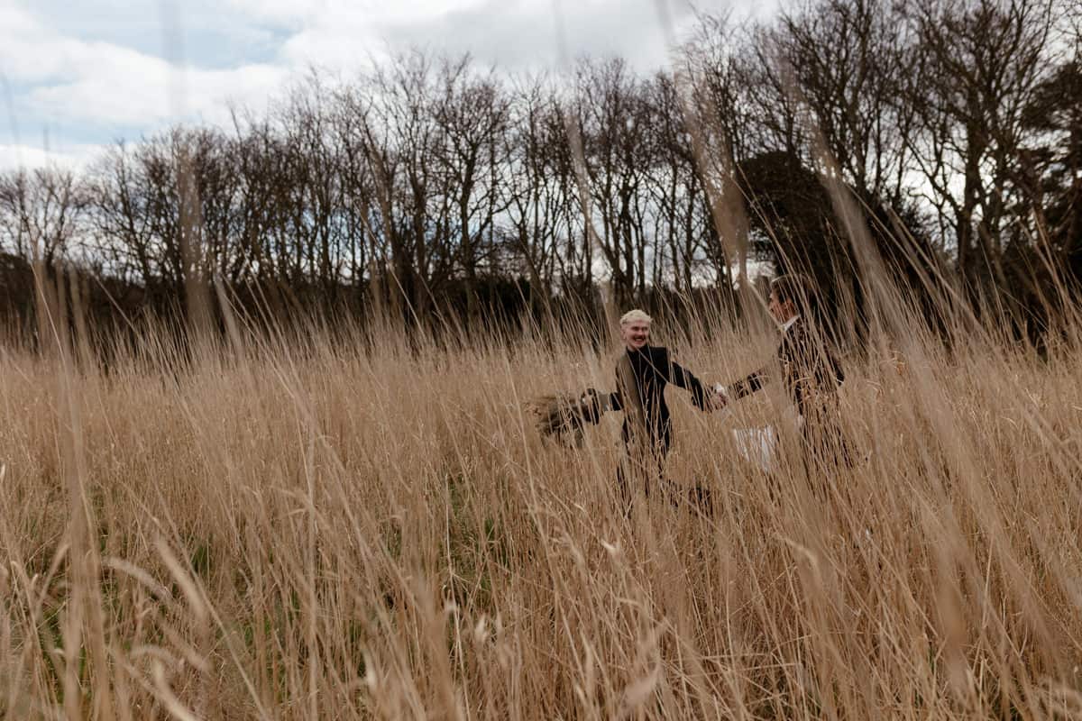 Two people running hand‑in‑hand through tall grass at the Dunglass Estate, framed by bare trees and an open sky.