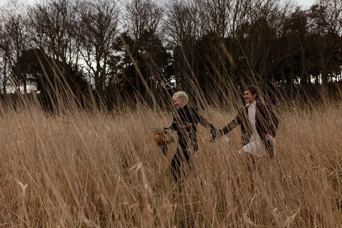 Two people running hand‑in‑hand through tall dry grass, one carrying flowers, with bare trees in the background.