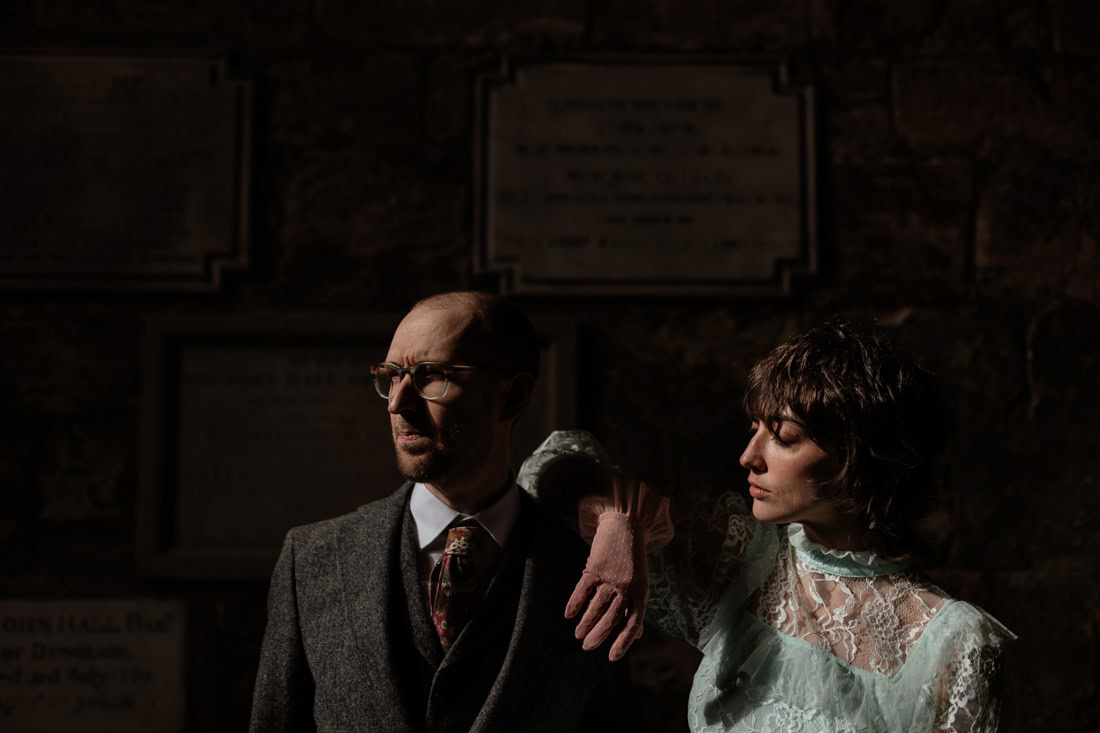 Two people standing in a dim stone interior, one in a suit and one in blue lace, lit softly against memorial plaques.