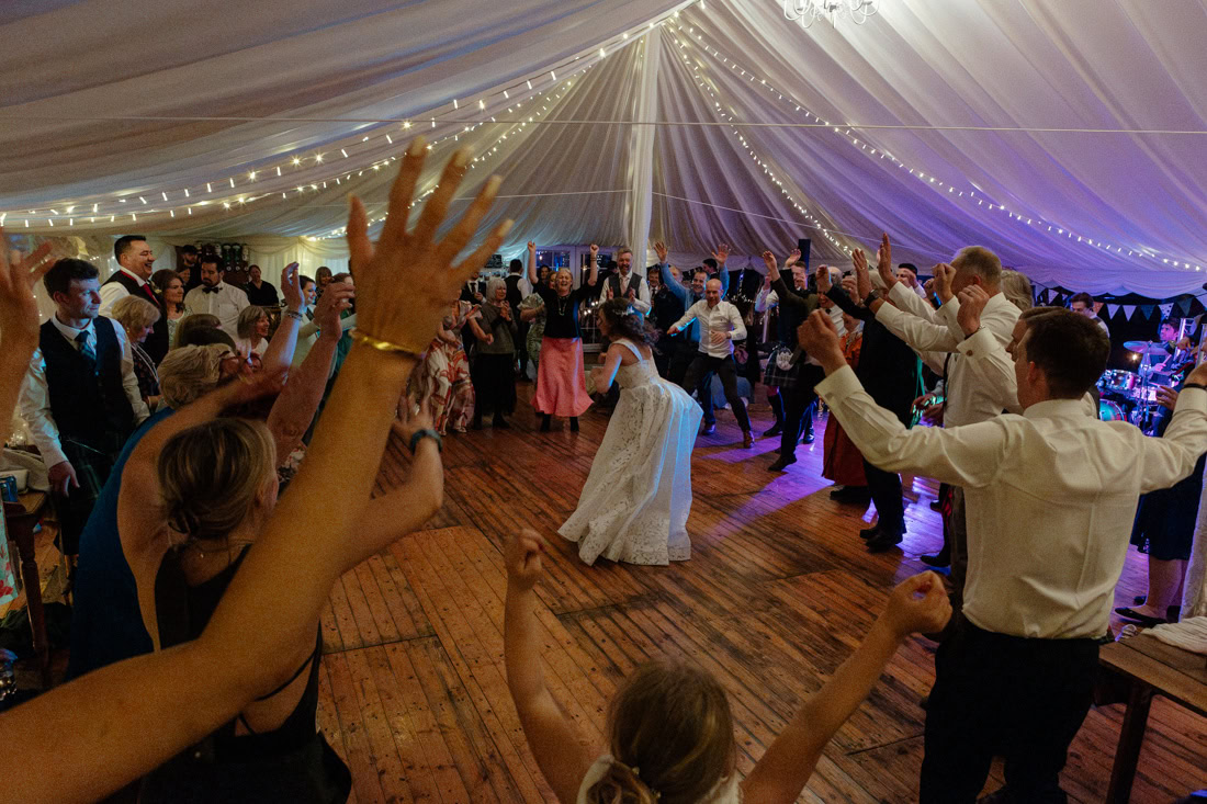 A lively wedding reception with the bride dancing in the centre as guests fill the fairy-lit marquee with hands raised in celebration.