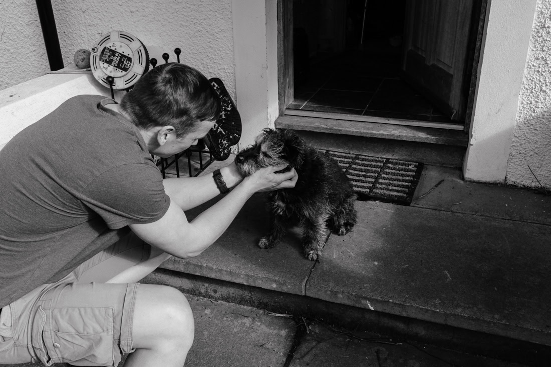 A man crouching on a doorstep to pet a small scruffy dog, captured in black and white .
