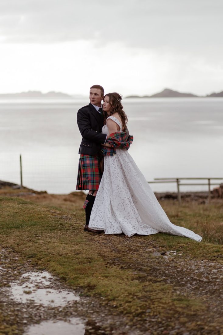 A bride and groom pose together on a grassy shore with a serene bay and distant hills in the background. The groom wears a traditional Scottish kilt in red tartan and a dark jacket, while the bride wears a white lace wedding dress and a matching tartan shawl. The overcast sky adds a romantic and moody atmosphere to the scene.