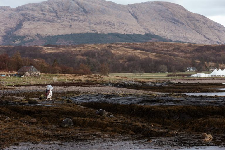 A tiny bride and groom walk along the rocky Cuil Bay shoreline with an umbrella, dwarfed by vast Scottish Highland mountains.