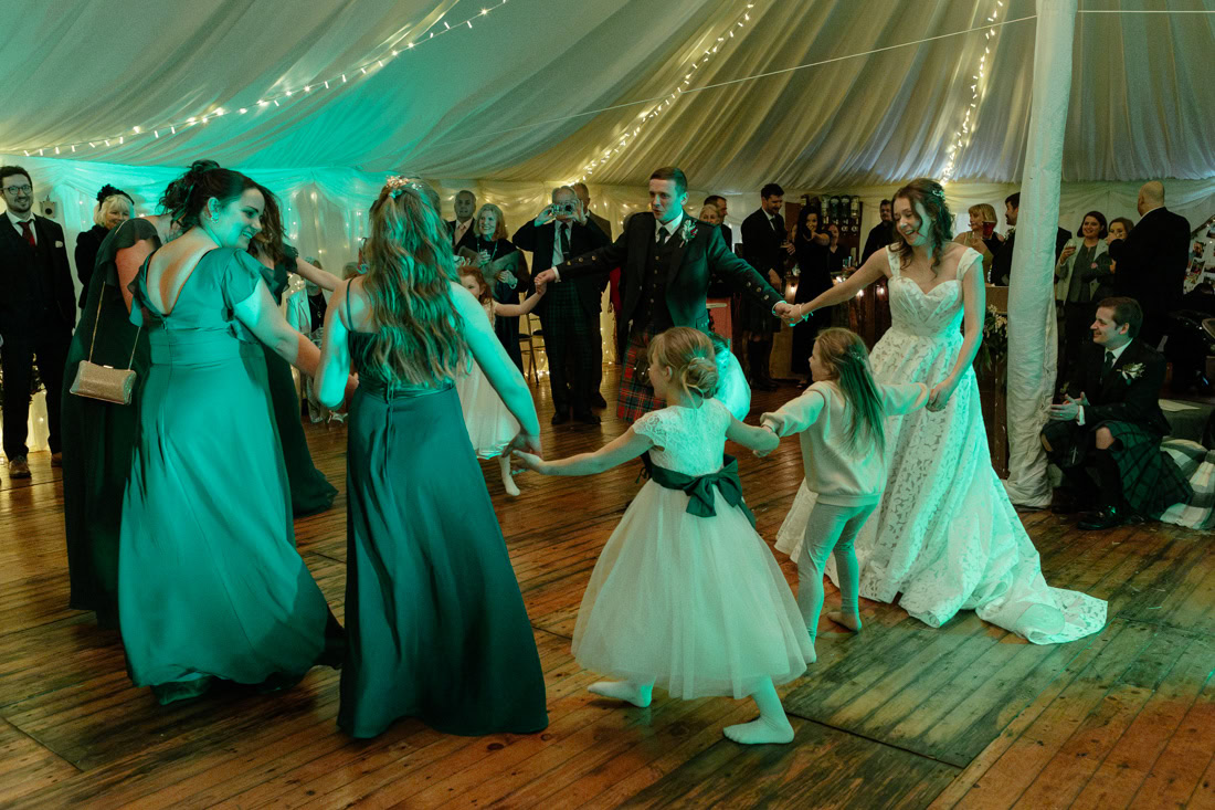 Bride and bridesmaids in green dresses dance joyfully with flower girls in a fairy-lit marquee bathed in green light.