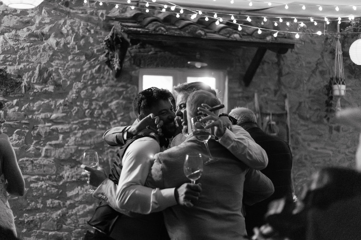Group hug under string lights; guests with wine glasses celebrate beside rustic stone wall.