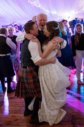 Bride and groom embrace with guest at lively Scottish beach wedding, surrounded by music, laughter, and celebration.