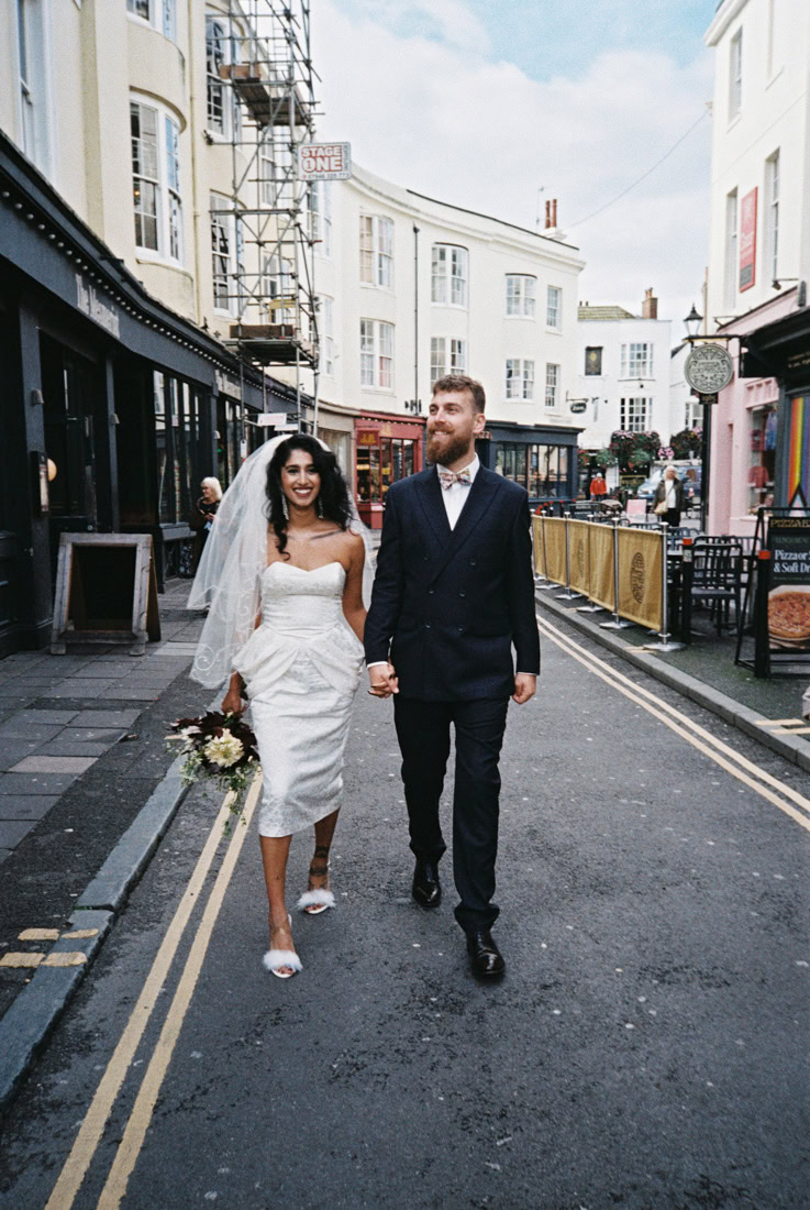 Bride and groom stroll past scaffold and cafés, bouquet in hand, celebrating in city sunshine; shot on 35mm film.