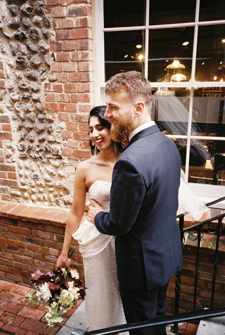 Couple embrace and smile by brick and stone wall, bride holding bouquet; shot on 35mm film.