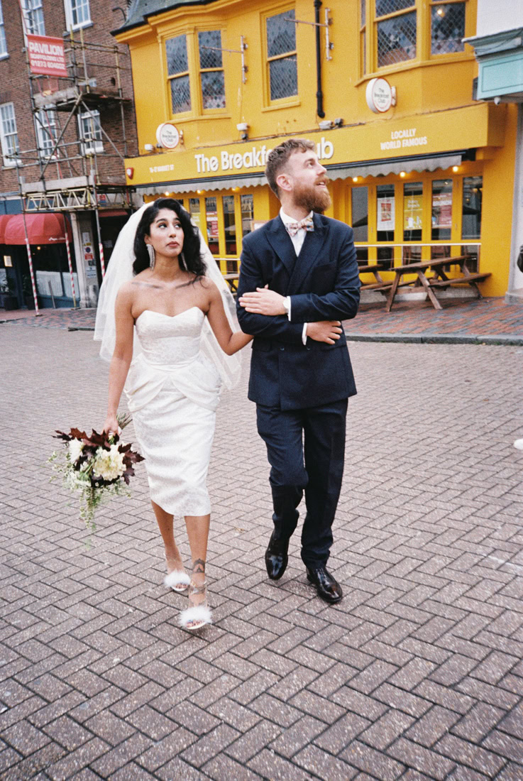 Couple walking past yellow café in wedding attire, smiling and arm-in-arm on a Brighton street.