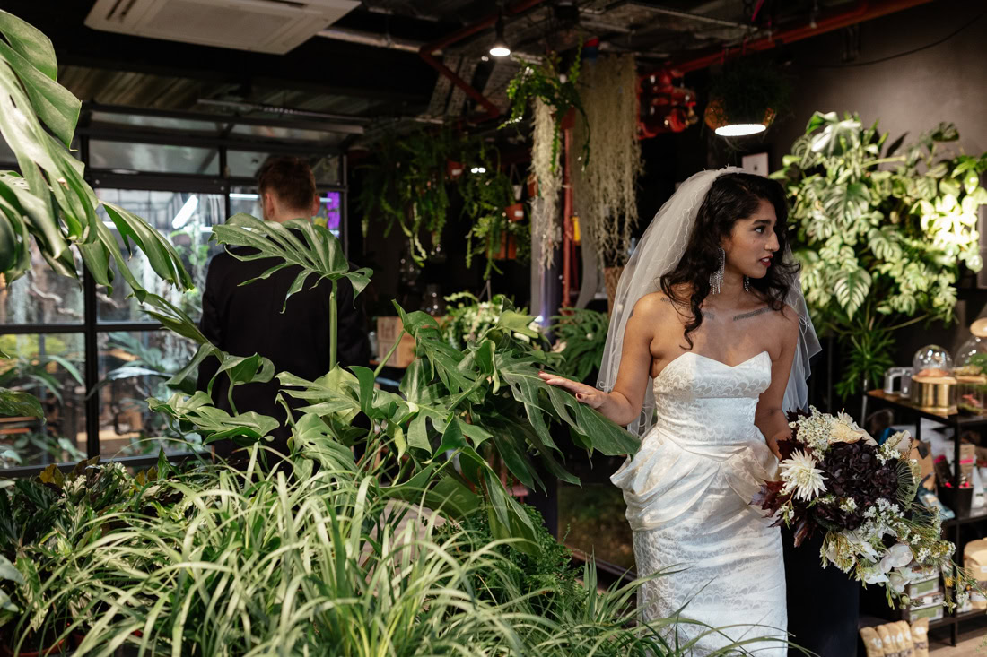 Bride in white gown holding bouquet amid lush indoor plants, groom visible in background.