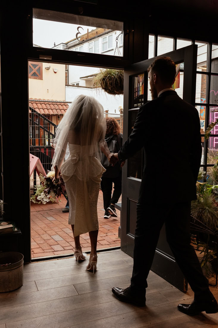 Newlyweds step from a doorway onto brick pavement; bride in veil and gown holding a bouquet.