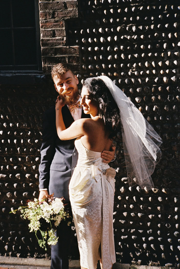 35mm image of a Couple stand close by brick wall, bouquet and veil framed in dramatic light