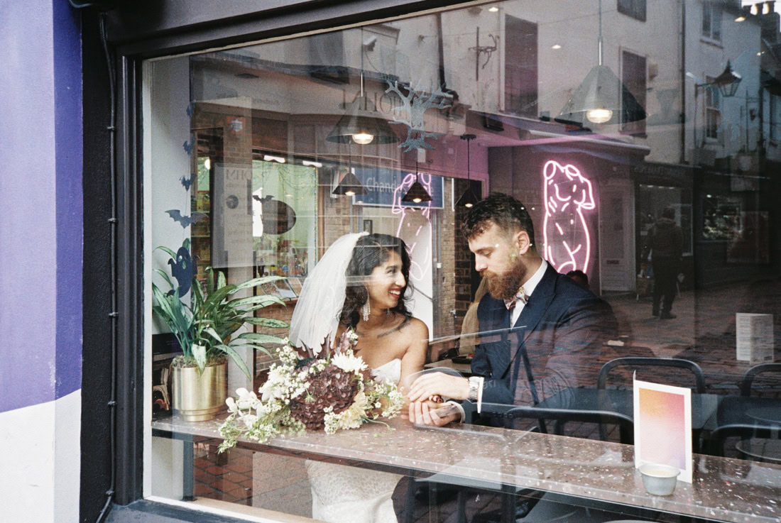 Couple sit inside café, bouquet on table, holding hands through window on wedding day.