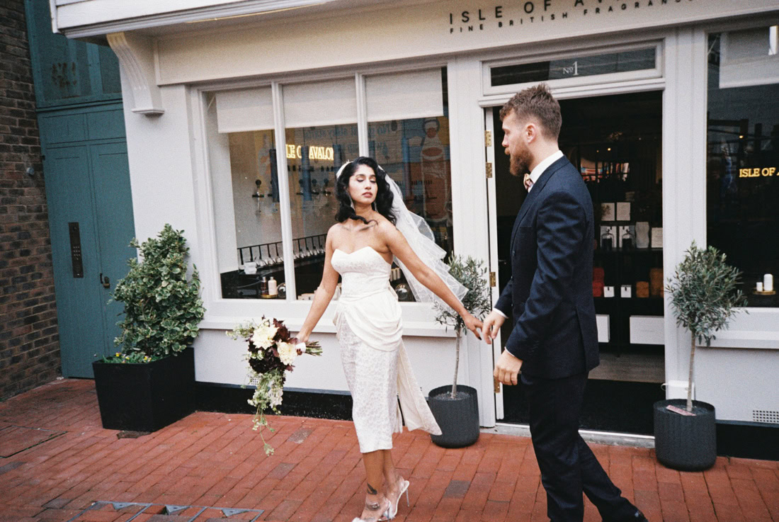 Bride leads groom past “Isle of Avalon” shop, bouquet in hand, shot on 35mm film