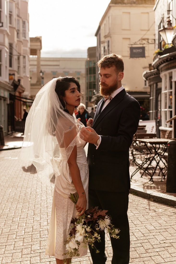 Groom adjusts bride’s veil on cobbled Brighton street, warm light and urban charm surround them.
