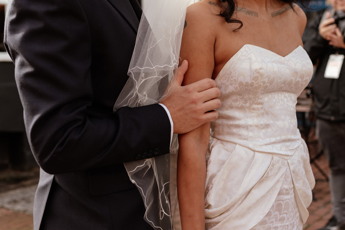Close-up of groom holding brides arm, bride’s textured gown and veil highlighted in outdoor setting.