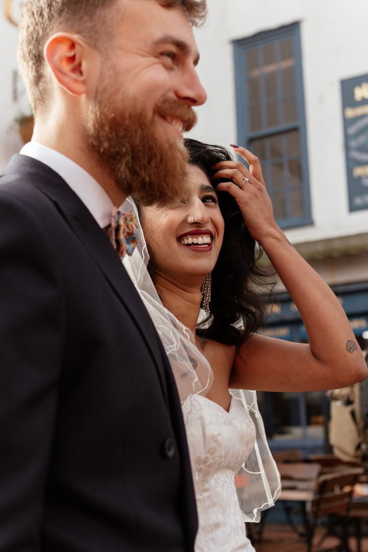 Bride smiles and adjusts hair beside groom in bow tie, candid moment in front of blue windows.