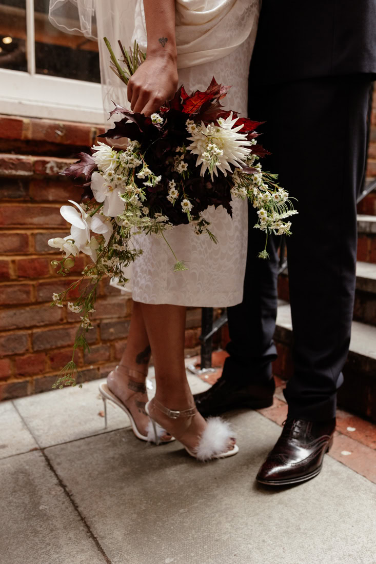 Close-up of couple’s shoes and large flower bouquet on stone pavement, bride in feathered heels