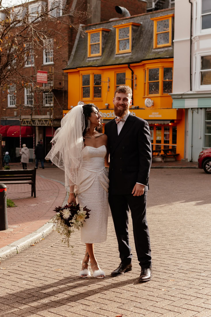 Bride and groom pose on brick street near orange storefront, bouquet in hand, joyful and elegant.