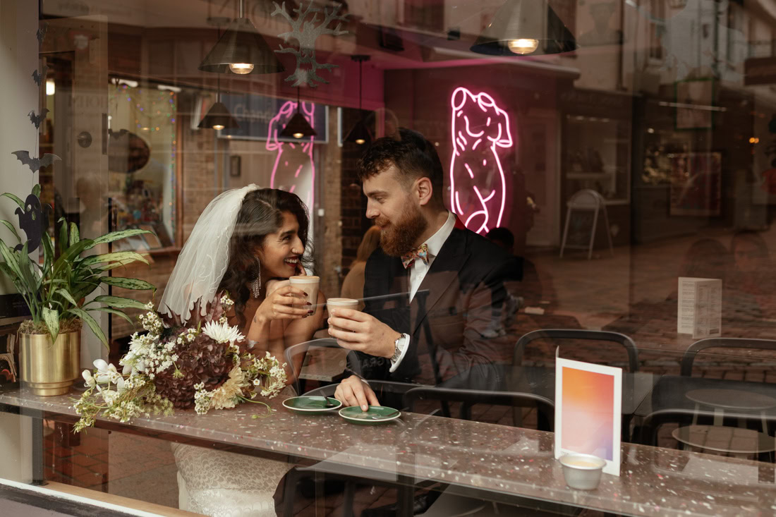 Couple sit at a Brighton café table, holding coffee cups, bouquet on table, seen through the window.