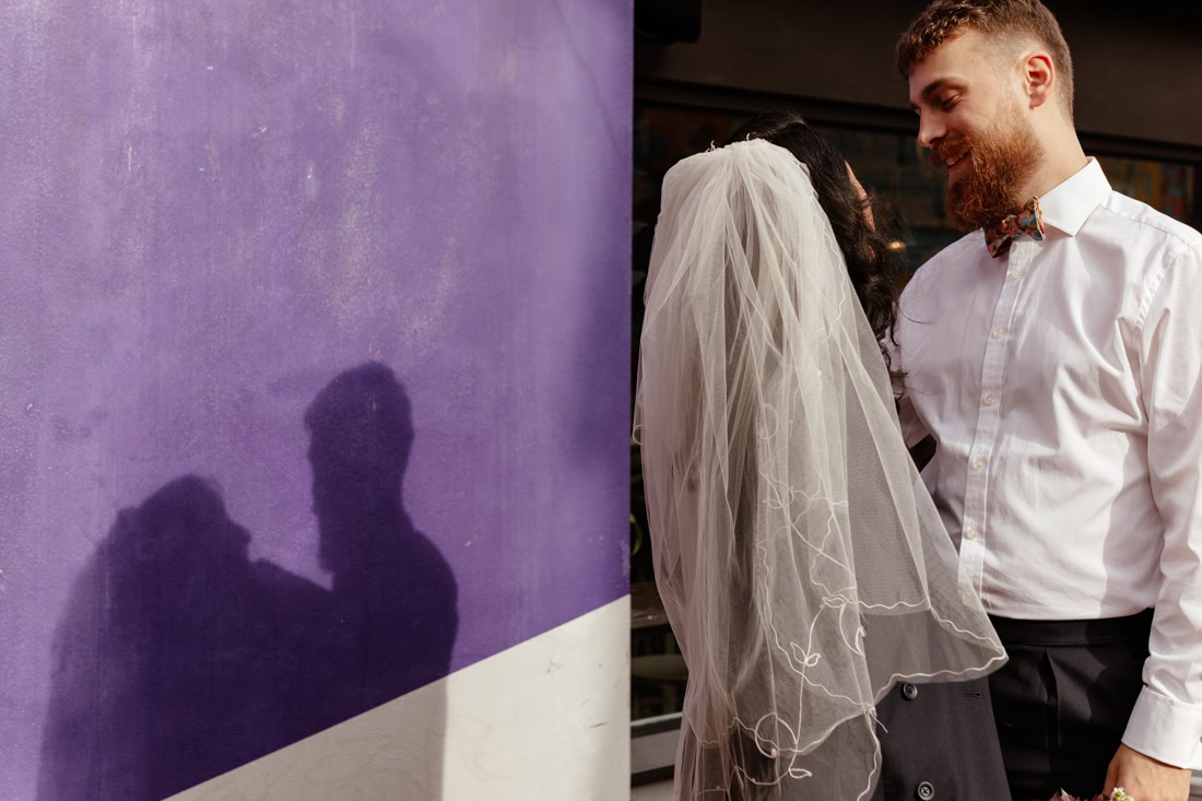 Couple pose against a textured stone wall, dramatic light highlighting veil and suit.