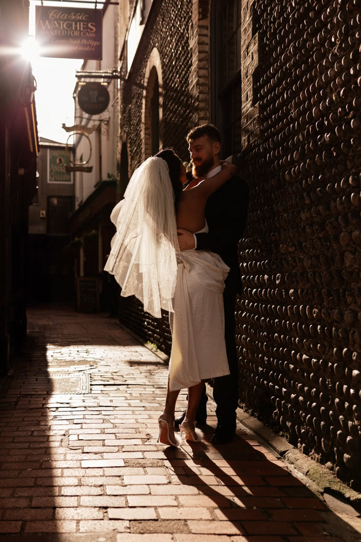 Groom hugs bride through sunlit narrow brick alley, shadows and shop signs framing them.