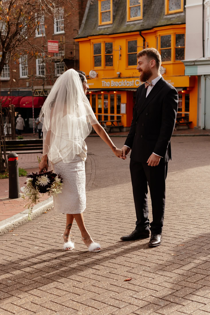 Couple in wedding dress and suit holding hands and smiling outside orange café, sharing a moment.