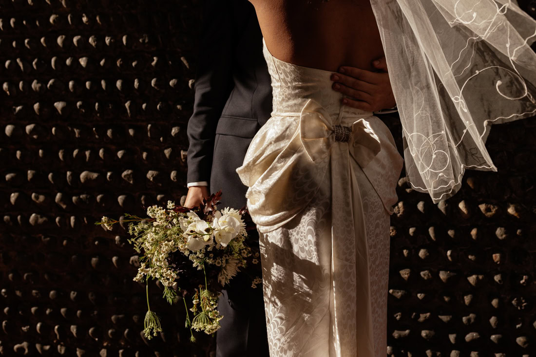 Close-up of couple with bouquet and veil, bride’s off-shoulder dress and bow in warm light.
