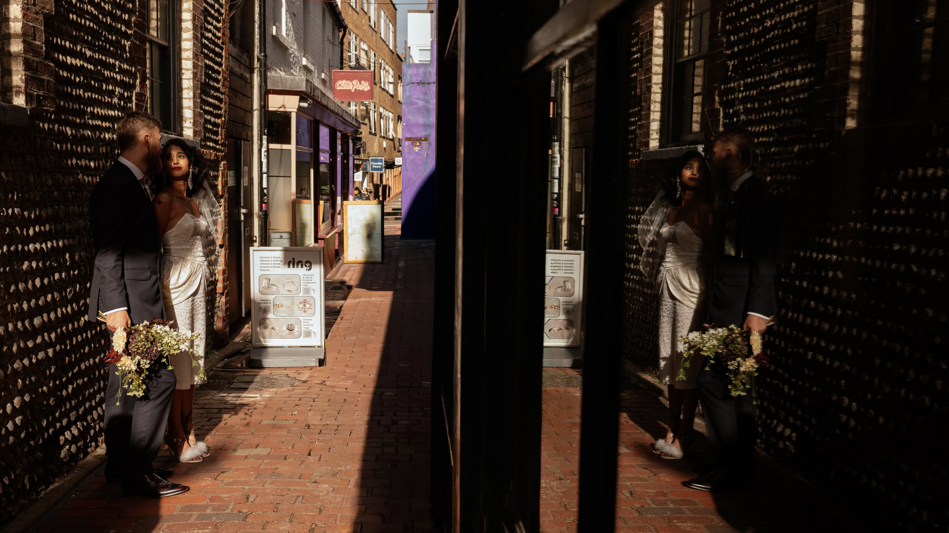 Couple in wedding attire stand in a narrow brick alley, bouquet in hand, reflection in a shop window