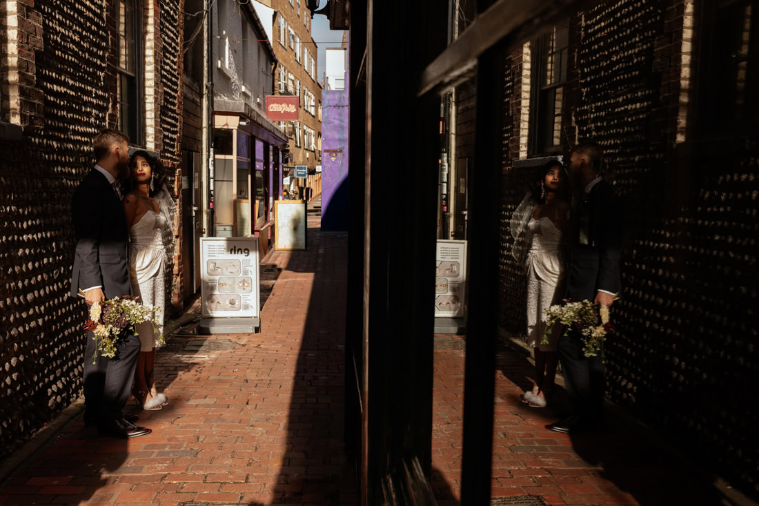 Couple in wedding attire stand in a narrow brick alley, bouquet in hand, reflection in a shop window