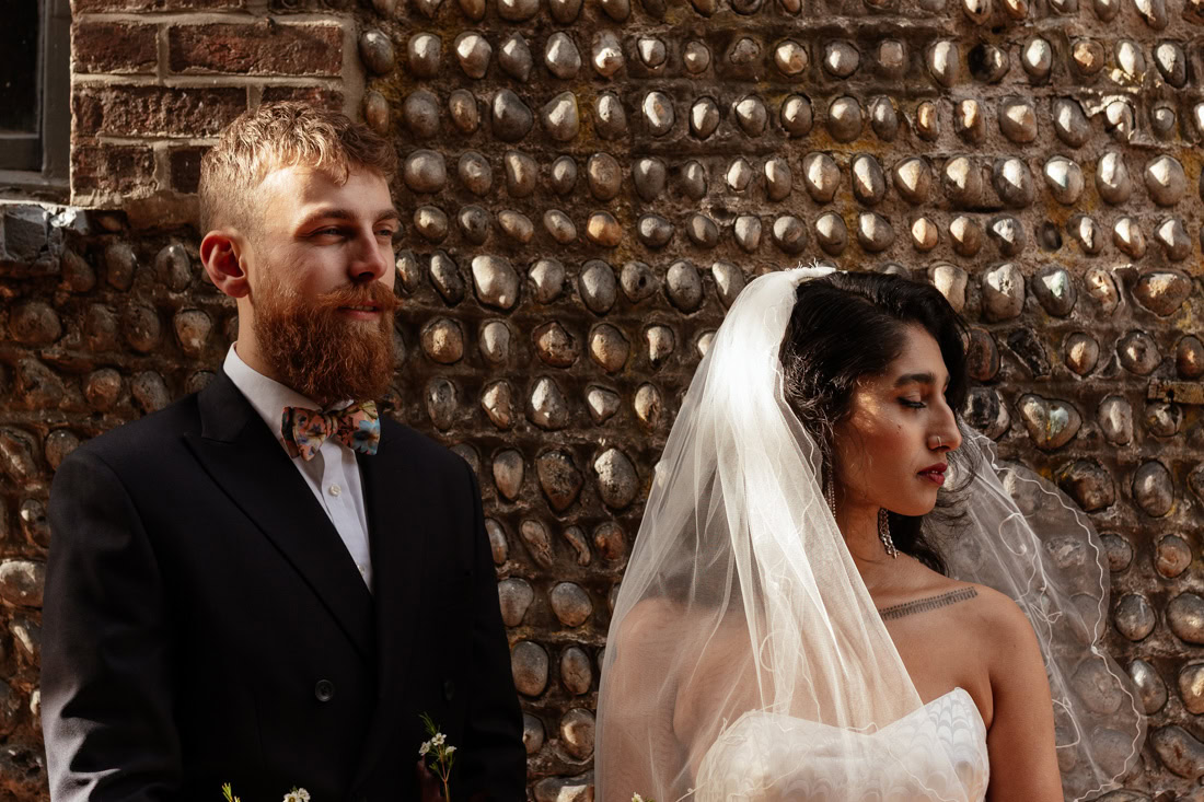 Couple stand close in wedding attire, dramatic light highlights dress, bow tie, and veil.