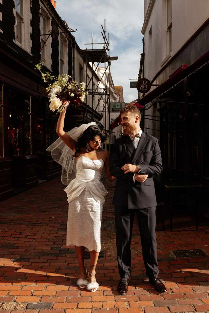 Bride and groom stand smiling arm-in-arm in a brick alley, bride holding bouquet aloft in warm light.