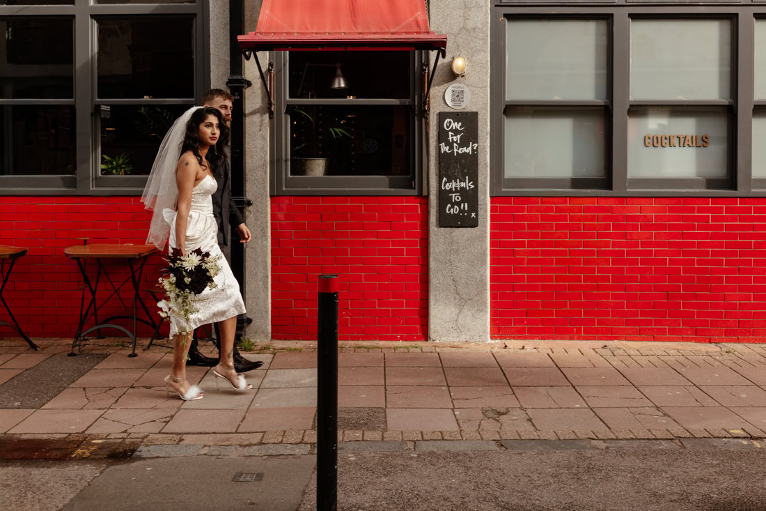 Bride and groom walk past a red-brick café with outdoor tables and a “Cocktails to go” sign.