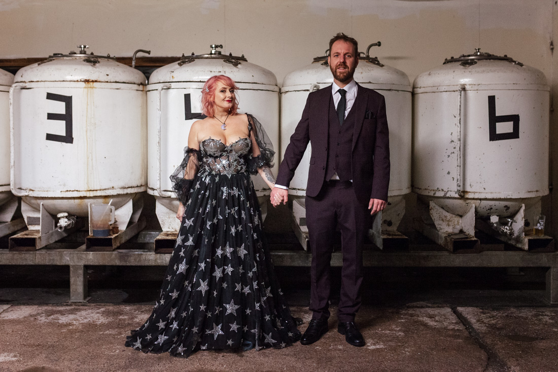 Two people; one in a starry black wedding gown with pink hair, the other in a suit, pose before large brewery tanks.