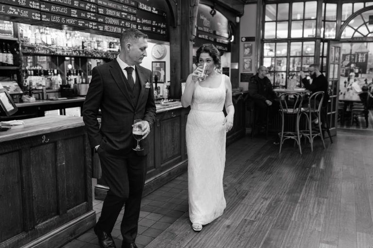 Bride and groom in wedding attire share drinks at the embankment pub bar in a relaxed black‑and‑white scene.