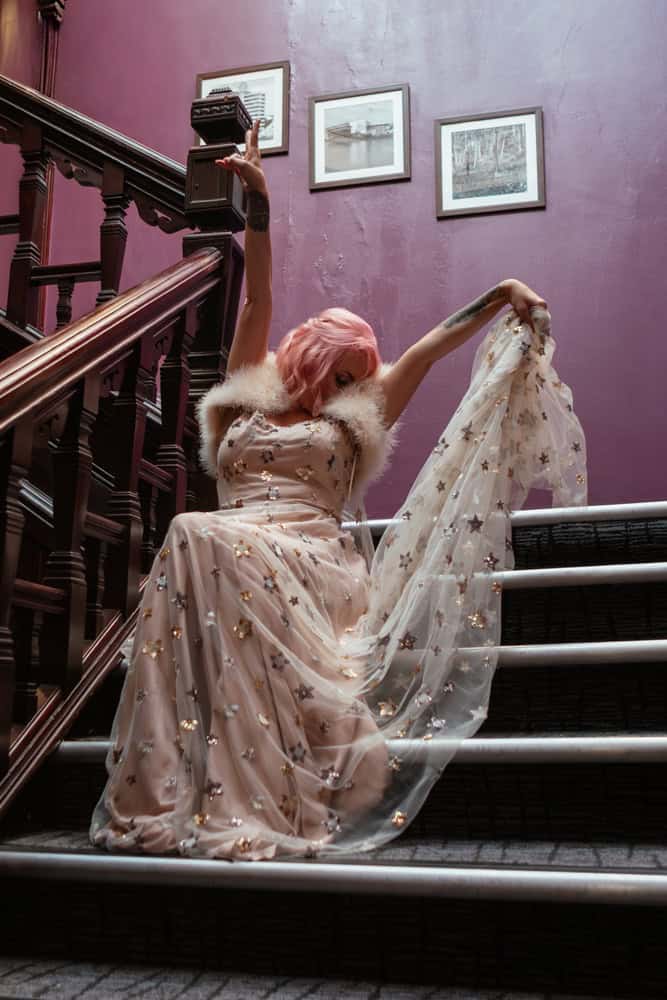 Pink-haired bride poses on purple stairwell in star-embellished gown and fur stole at micro wedding in Nottingham City.