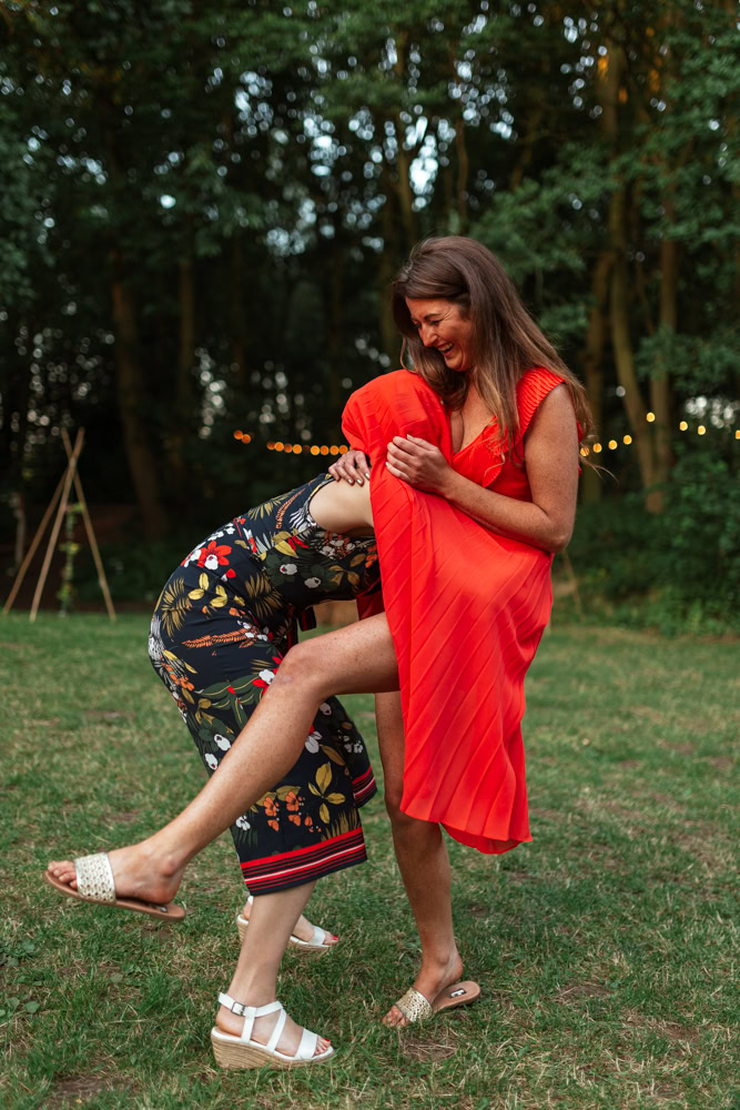 Two people laughing outdoors as one playfully ducks under the other’s red dress on a grassy lawn.