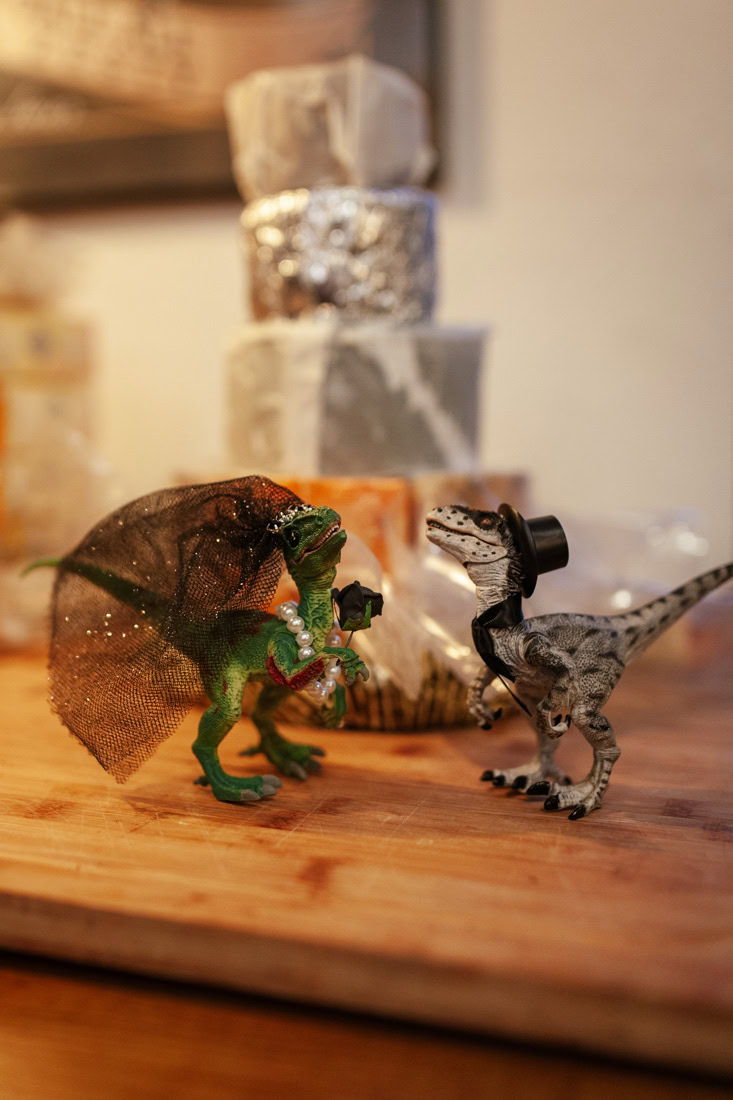 Two small dinosaur figurines dressed as a wedding couple stand before a stacked cheese cake